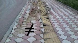 Görselde kiremit ve gri renkli, baklava dilimi şeklindeki döşeme taşlarından oluşan bir kaldırım bulunuyor. Kaldırımın sağ kenarında küçük bir yeşillik, sol yanında ise cadde bulunuyor. Kaldırımın ortasında bulunan sarı kabartmalı şeritler ve sağ tarafındaki döşeme taşları oldukça bozuk bir halde. Kaldırımın bozuk tarafında eşit değil sembolü yer alıyor.