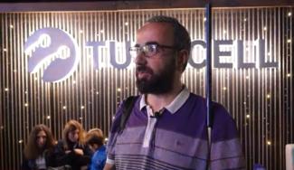 Cimri oyununa gelmiş yakası beyaz mor tshirtlü, siyah sakallı, gözlüklü ve beyaz bastonlu izleyici ayakta duruyor. Arka planda yer alan Turkcell yazılı ve logolu büyük ışıklı panonun sol köşesinde iki kadın ve bir erkek çocuk görülüyor.