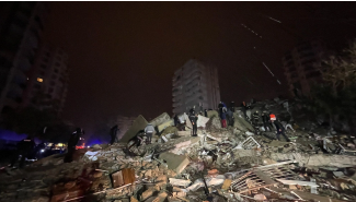 Karanlık bir deprem bölgesinde, üzerine ışık vurmuş enkaz yığınında dağınık bir şekilde birçok insan arama kurtarma çalışmaları yapıyor.
