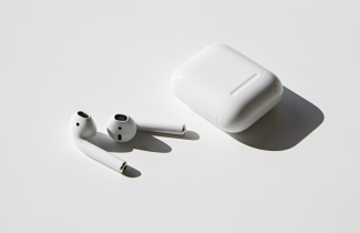 Beyaz bir zemin üzerinde dikdörtgen şeklinde beyaz airpod kutusu ve hemen yanında beyaz kablosuz kulaklıklar yer alıyor.