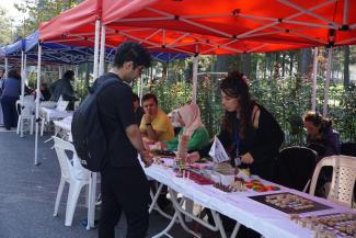 Beyaz Baston ve Erişilebilirlik Festivali’nin Oyun ve Hobi Standı. Beyaz örtülü masalar üzerinde Jenga, rubik küpü, satranç gibi oyunlar yer alıyor. Siyah uzun saçlı, siyah hırkalı görevli ayakta ve bir oyunun anlatımını yapıyor. Karşısında siyah sırt çantalı, gözlüklü bir erkek onu dinliyor. Arka planda sarı tişörtlü kör bir erkek ile yeşil bluzlü beyaz başörtülü kör bir kadın oturuyor.