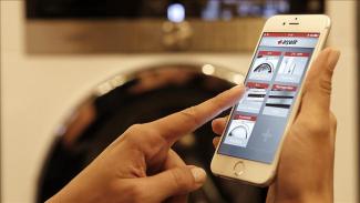 Arka planda belirsiz bir çamaşır makinesi, önde sadece ellerini gördüğümüz cep telefonu tutan bir kadın. Cep telefonunda Arçelik uygulaması var ve ekranda açık, pişirmeye hazır, duraklatıldı, 3°C -18°C gibi komutlar görülüyor. Kadının sol işaret parmağı açık komutu üzerinde.