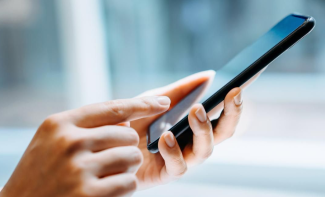 Yatay dikdörtgen formdaki görselde yandan görüntülenmiş bir çift kadın eli. Elin biri telefonu tutuyor, diğer elin işaret parmağı telefonun ekranına dokunuyor.