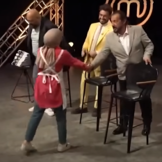 Master Chef programından bir sahne. Yazıda ismi geçen Melek Hanım üç jürinin ortak oyuyla kabul aldığı için kendisine giydirilmiş olan beyaz şef önlüğüyle kameraya arkası dönük bir şekilde Mehmet Şef’le tokalaşıyor. Dizinin biraz üzerinde kırmızı gömlek, mavi kot pantolon ve spor ayakkabı giymiş ve başörtüsü varmış. Mehmet Şef’in hemen yanında sarı ceketi ile ayakta duran Danilo gülüyor. Onun yanında ise Soner Şef taburesine otururken görülüyor.
