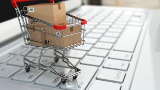 Görselde yakından görüntülenmiş bir laptop klavyesi, üzerinde minimal bir metal alışveriş sepeti yer alıyor. Alışveriş sepetinin içerisinde, üstleri bantlanmış ve barkodları yapıştırılmış üç adet koli bulunuyor.
