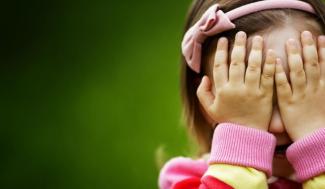 Arka planında yeşil fon olan görselin sağ tarafında küçük, kumral bir kız çocuğu ellerini yüzüne kapatmış bir şekilde duruyor. Çocuğun omuzlarına kadar olan saçlarına taktığı pembe renkli tacında aynı renkte bir fiyonk bulunuyor. Göğüsten üstü görünen bluzu pembe ve sarı renklerde.
